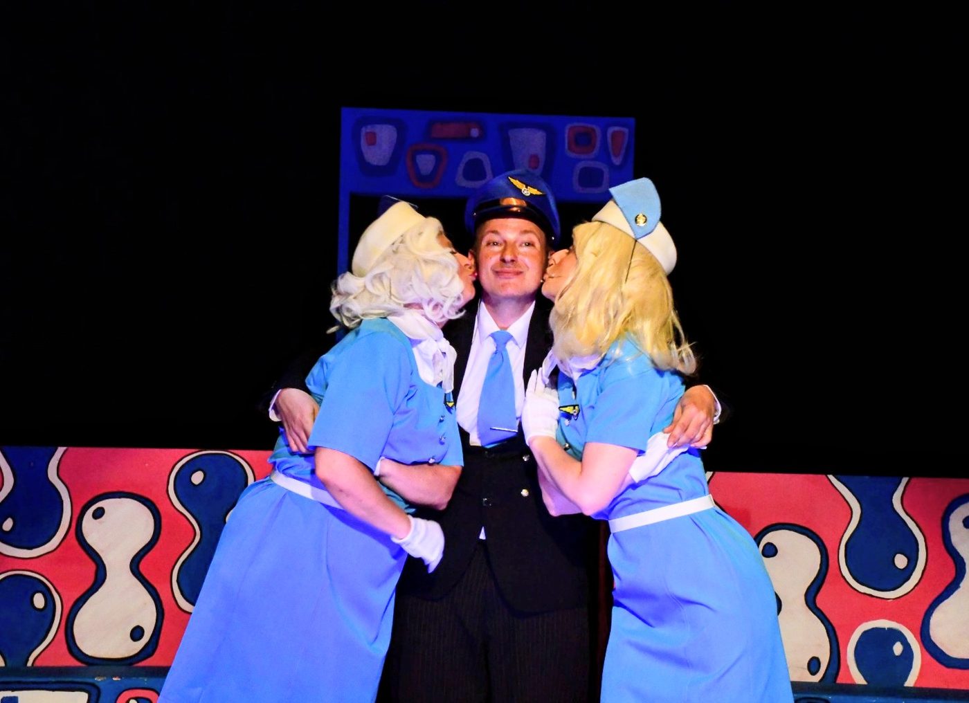 A pilot in uniform grins as two air stewardesses in blue plant a kiss on each cheek