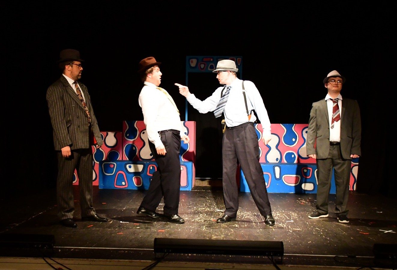 Four suited detectives face off mid-scene against a pop-art set